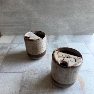 May include: Two ceramic mugs with a speckled beige and brown glaze. Each mug has a dark brown band near the base and a unique, angled opening. The mugs are placed on a light gray tiled surface.