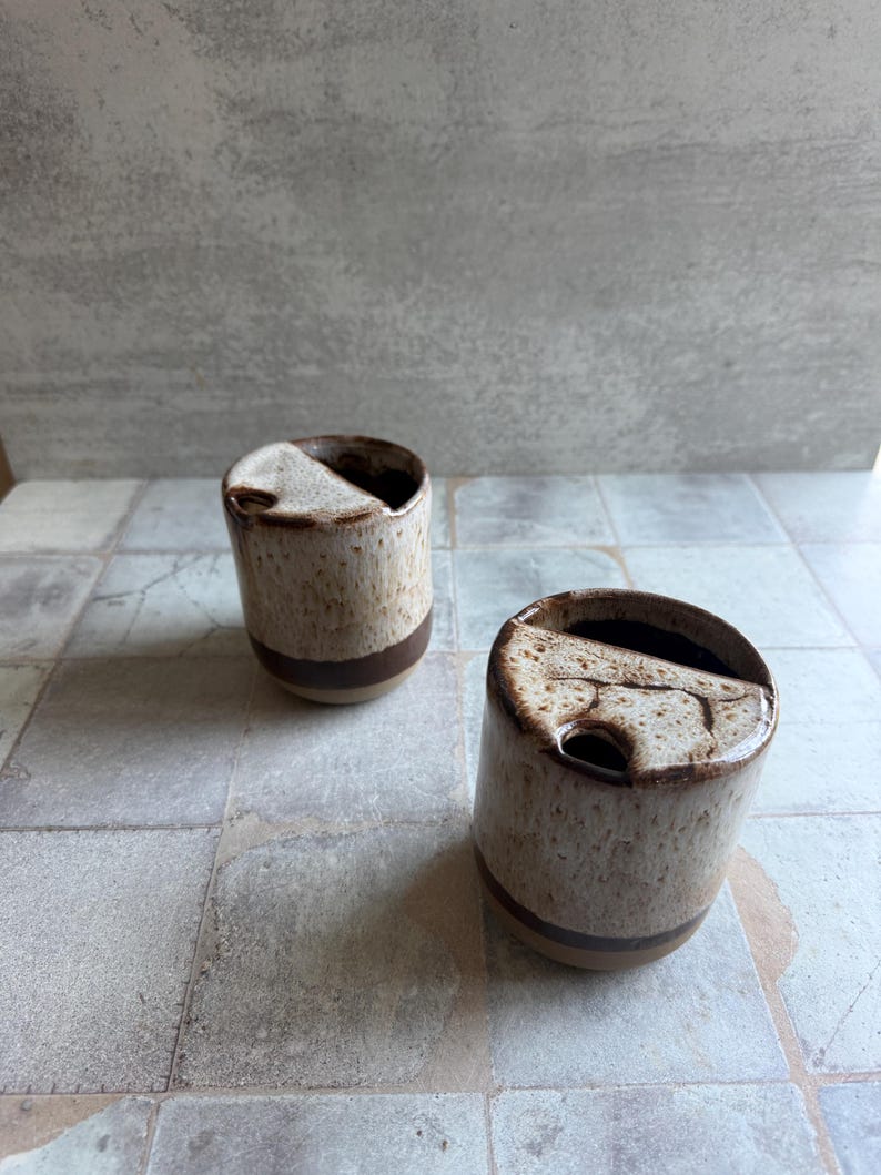 May include: Two ceramic mugs with a speckled beige and brown glaze. Each mug has a dark brown band near the base and a unique, angled opening. The mugs are placed on a light gray tiled surface.