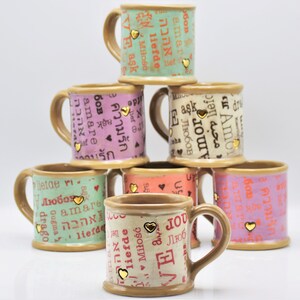 May include: A stack of ceramic mugs in various colors, including green, pink, and peach. Each mug features the word "love" in multiple languages, along with gold heart accents. The mugs have brown handles and rims.