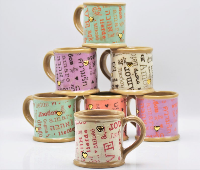 May include: A stack of ceramic mugs in various colors, including green, pink, and peach. Each mug features the word "love" in multiple languages, along with gold heart accents. The mugs have brown handles and rims.