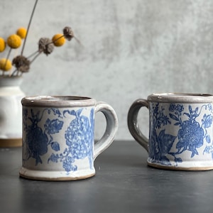 May include: Two ceramic mugs with blue floral patterns. The mugs have a light gray handle and rim, and a white base. The mugs are displayed on a dark surface, with a vase of yellow flowers in the background.