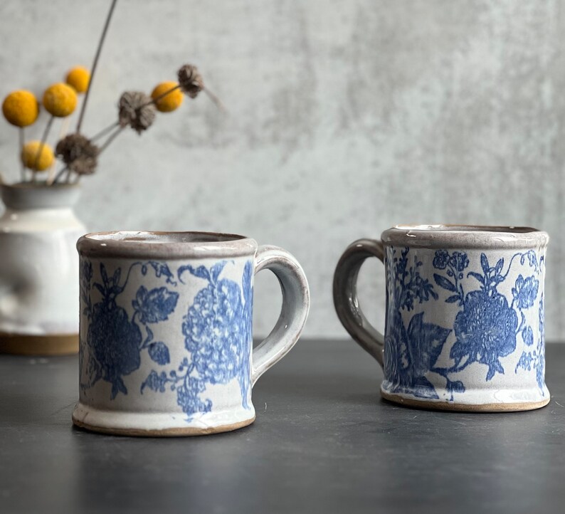 May include: Two ceramic mugs with blue floral patterns. The mugs have a light gray handle and rim, and a white base. The mugs are displayed on a dark surface, with a vase of yellow flowers in the background.