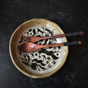 Puede incluir: Un plato de cer&aacute;mica con un dise&ntilde;o en blanco y negro de p&aacute;jaros y animales. El plato est&aacute; sobre una superficie negra y tiene dos cucharas de madera encima.