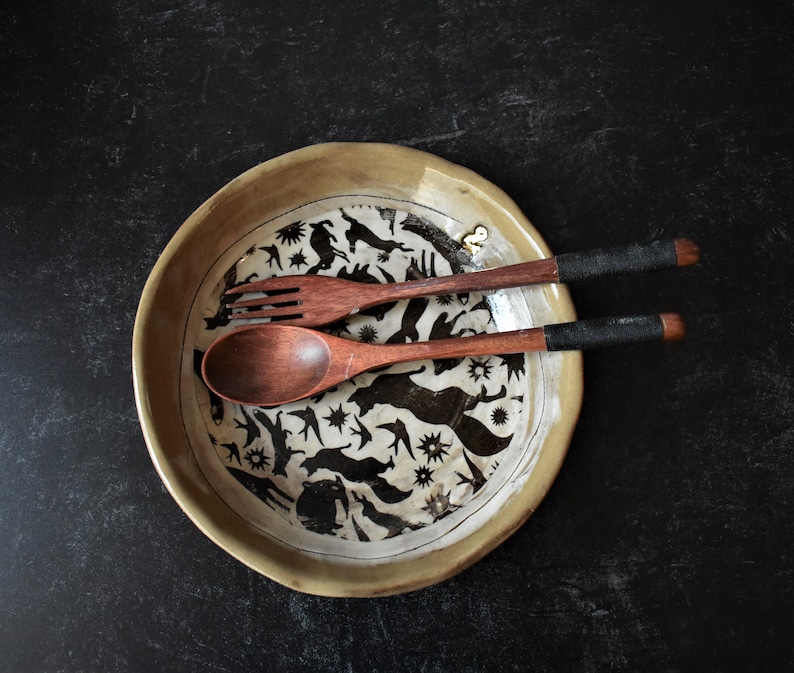 Puede incluir: Un plato de cer&aacute;mica con un dise&ntilde;o en blanco y negro de p&aacute;jaros y animales. El plato est&aacute; sobre una superficie negra y tiene dos cucharas de madera encima.