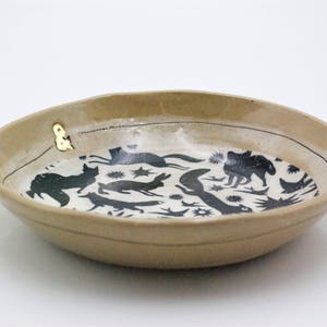 May include: A beige ceramic bowl with a black and white design of animals and stars. The bowl has a gold accent on the rim.
