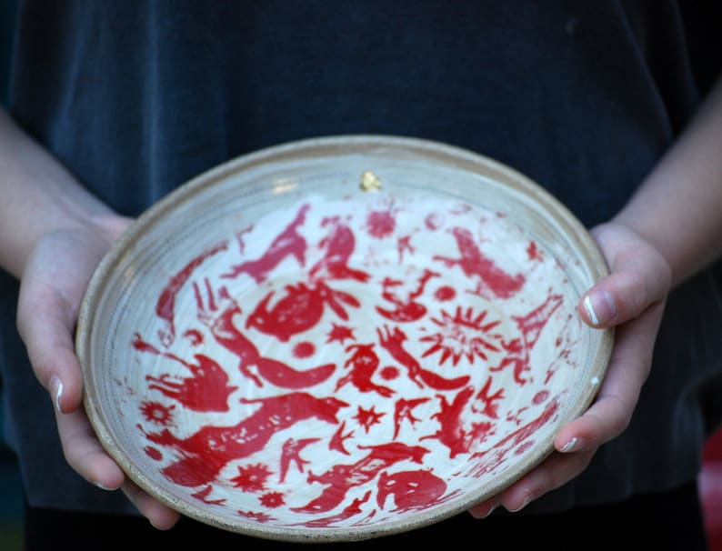 May include: A white ceramic bowl with a red and gold design. The bowl is decorated with a variety of stylized animal and plant motifs.