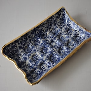 Puede incluir: Bandeja rectangular de cer&aacute;mica con un estampado floral azul y borde dorado. La bandeja tiene un borde ligeramente ondulado y una serie de protuberancias redondeadas en el interior. El dise&ntilde;o floral es un patr&oacute;n cl&aacute;sico azul y blanco.