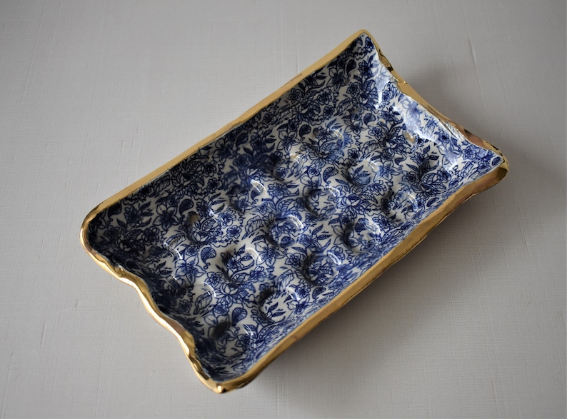 Puede incluir: Bandeja rectangular de cer&aacute;mica con un estampado floral azul y borde dorado. La bandeja tiene un borde ligeramente ondulado y una serie de protuberancias redondeadas en el interior. El dise&ntilde;o floral es un patr&oacute;n cl&aacute;sico azul y blanco.