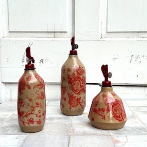 May include: Three ceramic bottles with red floral patterns and red metal tops. The bottles are different sizes and shapes. The bottles are on a white tiled surface.