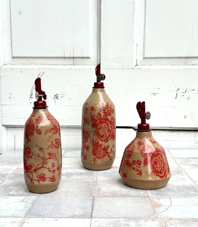 May include: Three ceramic bottles with red floral patterns and red metal tops. The bottles are different sizes and shapes. The bottles are on a white tiled surface.