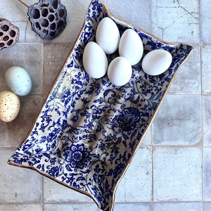 Puede incluir: Una fuente rectangular de cer&aacute;mica con un estampado floral azul y ribete dorado, que contiene cinco huevos blancos. La fuente est&aacute; colocada sobre una superficie embaldosada, con vainas de semillas secas y huevos decorativos cerca.