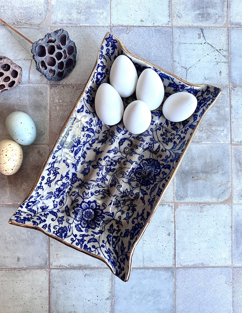 Puede incluir: Una fuente rectangular de cer&aacute;mica con un estampado floral azul y ribete dorado, que contiene cinco huevos blancos. La fuente est&aacute; colocada sobre una superficie embaldosada, con vainas de semillas secas y huevos decorativos cerca.