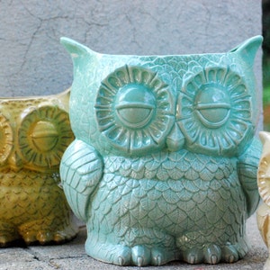 May include: A selection of ceramic owl-shaped planters in different colours. The main planter is a light turquoise, with detailed feather and eye designs. Other planters are visible in shades of yellow and beige. Suitable for indoor or outdoor use, these planters are decorative.