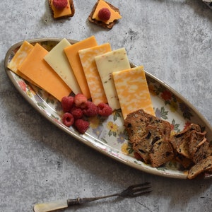 K&ouml;nnte beinhalten: Eine ovale Servierplatte mit floralem Muster, auf der verschiedene K&auml;sesorten, Cracker und Himbeeren liegen.