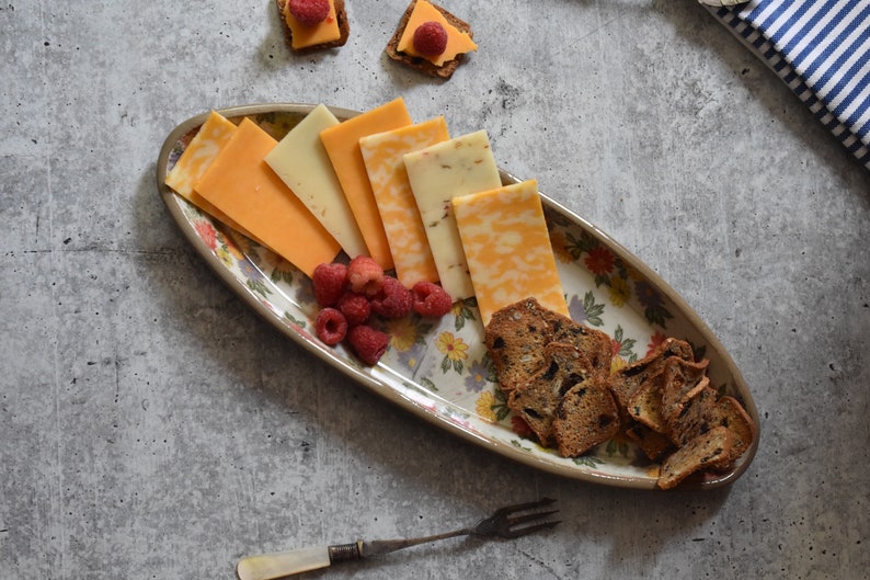 K&ouml;nnte beinhalten: Eine ovale Servierplatte mit floralem Muster, auf der verschiedene K&auml;sesorten, Cracker und Himbeeren liegen.