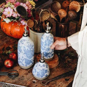 May include: Three ceramic oil and vinegar dispensers featuring a white base with a blue floral pattern and a silver metal pour spout. The set includes two tall cylindrical bottles and one shorter, rounded bottle, all with a smooth finish.