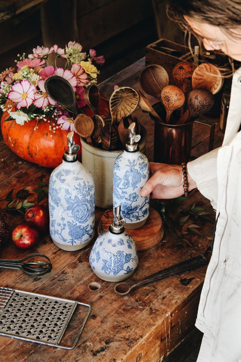 May include: Three ceramic oil and vinegar dispensers featuring a white base with a blue floral pattern and a silver metal pour spout. The set includes two tall cylindrical bottles and one shorter, rounded bottle, all with a smooth finish.