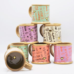 May include: A pyramid of ceramic mugs in various colors, including green, pink, and orange. Each mug features the word "love" in different languages, along with a small gold heart. The mugs have a brown handle and base.