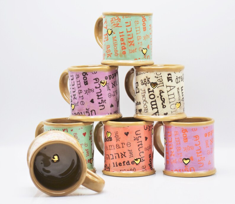 May include: A pyramid of ceramic mugs in various colors, including green, pink, and orange. Each mug features the word "love" in different languages, along with a small gold heart. The mugs have a brown handle and base.