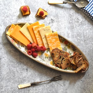 K&ouml;nnte beinhalten: Eine wei&szlig;e ovale Platte mit Blumenmuster zeigt eine Auswahl an geschnittenem K&auml;se und Crackern. Die Platte befindet sich auf einer grauen Oberfl&auml;che mit einer blau-wei&szlig; gestreiften Serviette und einem silbernen Buttermesser.