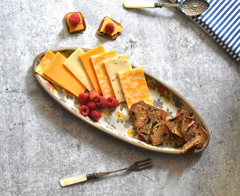 K&ouml;nnte beinhalten: Eine wei&szlig;e ovale Platte mit Blumenmuster zeigt eine Auswahl an geschnittenem K&auml;se und Crackern. Die Platte befindet sich auf einer grauen Oberfl&auml;che mit einer blau-wei&szlig; gestreiften Serviette und einem silbernen Buttermesser.