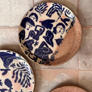 May include: A set of ceramic plates with a unique design. The plates feature a cream-colored background with navy blue illustrations of figures and foliage. The rims are a speckled pinkish-brown color, adding a touch of warmth to the design.