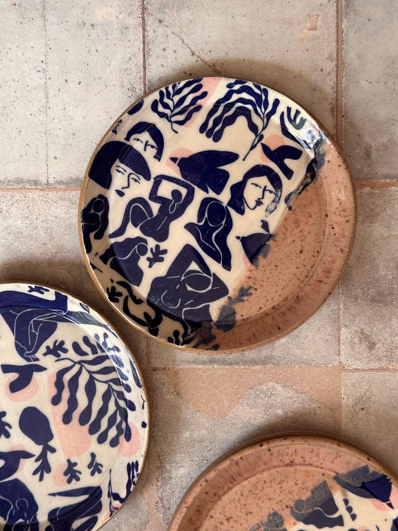 May include: A set of ceramic plates with a unique design. The plates feature a cream-colored background with navy blue illustrations of figures and foliage. The rims are a speckled pinkish-brown color, adding a touch of warmth to the design.