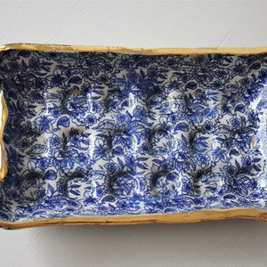 Puede incluir: Jabonera rectangular de cer&aacute;mica con un estampado floral azul sobre fondo blanco. Los bordes est&aacute;n adornados con dorado. La jabonera tiene una serie de protuberancias elevadas para el jab&oacute;n.