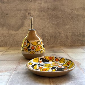 May include: A ceramic oil bottle and small dish set. The bottle has a brown base and a white top with a yellow, orange, and black pattern. The dish has a similar design. The set is on a tiled surface.
