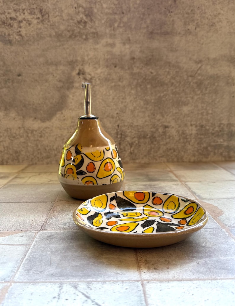 May include: A ceramic oil bottle and small dish set. The bottle has a brown base and a white top with a yellow, orange, and black pattern. The dish has a similar design. The set is on a tiled surface.
