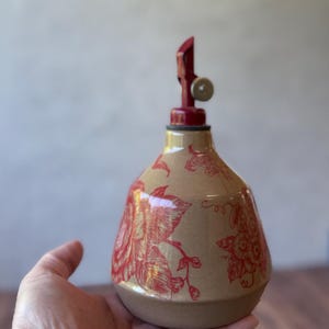 May include: A ceramic bottle with a red floral design and a red metal pump top. The bottle is hand-painted with a floral pattern and has a rustic, vintage look.