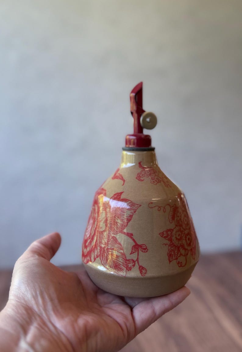 May include: A ceramic bottle with a red floral design and a red metal pump top. The bottle is hand-painted with a floral pattern and has a rustic, vintage look.