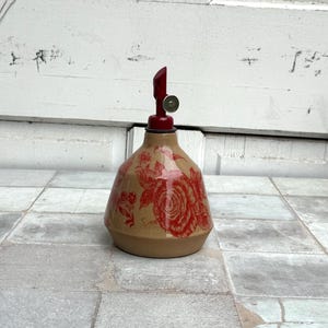 May include: A ceramic oil and vinegar bottle with a red floral design and a red and silver top.