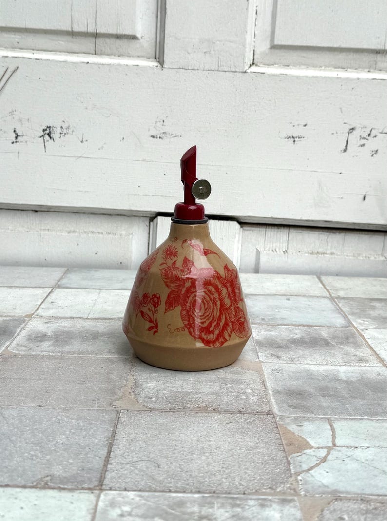 May include: A ceramic oil and vinegar bottle with a red floral design and a red and silver top.