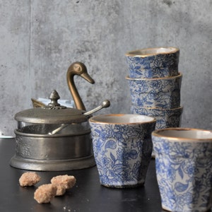 May include: A set of four blue and white ceramic cups with gold trim, stacked on top of each other. The cups have a floral pattern and are sitting on a black surface. There is a silver sugar bowl with a swan-shaped lid and a spoon in the background.