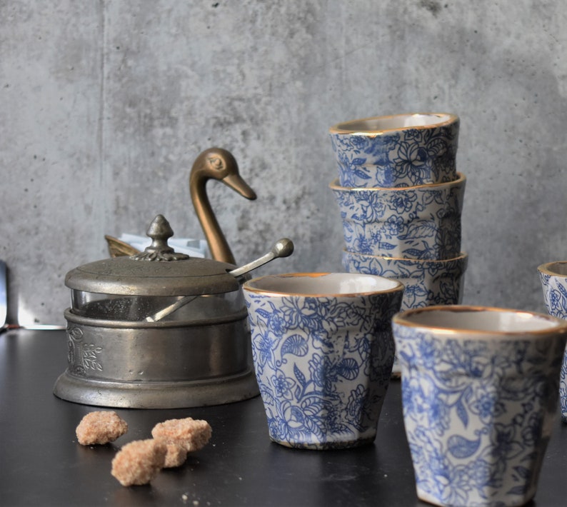 May include: A set of four blue and white ceramic cups with gold trim, stacked on top of each other. The cups have a floral pattern and are sitting on a black surface. There is a silver sugar bowl with a swan-shaped lid and a spoon in the background.