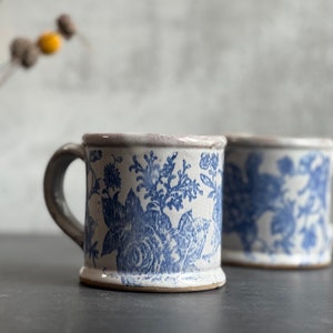 May include: Two ceramic mugs with blue floral patterns. The mugs have a white base, blue floral design, and gray handle. The mugs are on a dark surface with a blurred background. The mugs are a light beige color.