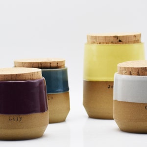 May include: Four ceramic jars with cork lids. The jars are in shades of purple, teal, yellow, and white, with a natural clay base. Each jar has a name printed on it: Lily, Jo, Je, and Callie.