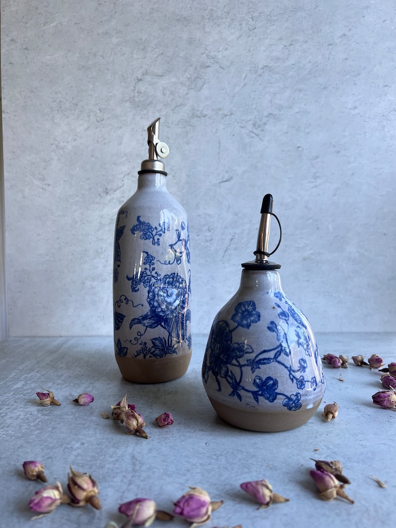 Two ceramic oil and vinegar bottles with blue and white floral designs. The bottles have a spout and a lid. The bottles are sitting on a light gray surface with dried rose petals scattered around them.