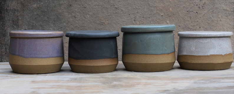 Peut inclure: Quatre pots en c&eacute;ramique avec couvercles. Chaque pot pr&eacute;sente une base marron clair et des sections sup&eacute;rieures de couleurs violet, noir, vert d'eau et blanc. Les couvercles reprennent la couleur sup&eacute;rieure de chaque pot.