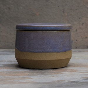 Peut inclure: Un pot en c&eacute;ramique fait &agrave; la main avec un couvercle. Le pot pr&eacute;sente un design bicolore, avec une section sup&eacute;rieure violet fonc&eacute; et une section inf&eacute;rieure beige. Le couvercle est de la m&ecirc;me couleur violet fonc&eacute;. Le pot est sur une surface en bois.