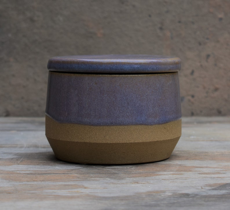 Peut inclure: Un pot en c&eacute;ramique fait &agrave; la main avec un couvercle. Le pot pr&eacute;sente un design bicolore, avec une section sup&eacute;rieure violet fonc&eacute; et une section inf&eacute;rieure beige. Le couvercle est de la m&ecirc;me couleur violet fonc&eacute;. Le pot est sur une surface en bois.