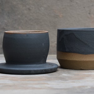 Peut inclure: Deux r&eacute;cipients en c&eacute;ramique faits &agrave; la main. L'un est de forme arrondie gris fonc&eacute; avec une large base. L'autre est un cylindre gris fonc&eacute; avec une bande marron en bas. Les deux ont une surface textur&eacute;e et une esth&eacute;tique rustique.