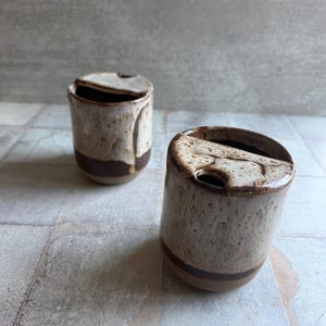May include: Two ceramic mugs with lids. The mugs are cylindrical with a speckled beige glaze and a dark brown band near the base. The lids are the same speckled beige color. The mugs are on a tiled surface.