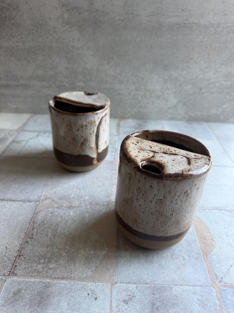 May include: Two ceramic mugs with lids. The mugs are cylindrical with a speckled beige glaze and a dark brown band near the base. The lids are the same speckled beige color. The mugs are on a tiled surface.
