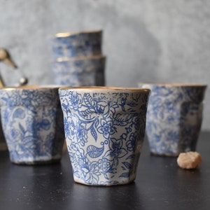May include: Set of five white ceramic cups with a blue floral pattern and gold trim. The cups are arranged in a row on a black surface.