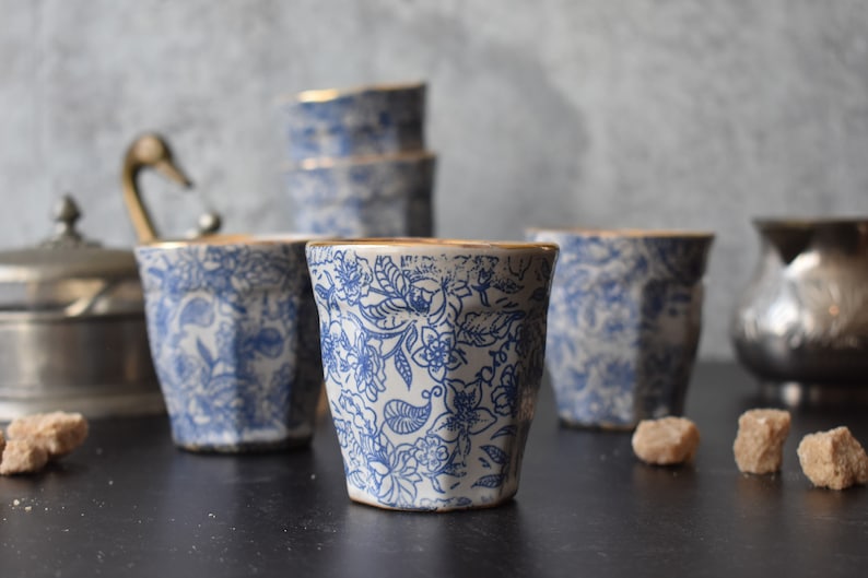 May include: Set of five white ceramic cups with a blue floral pattern and gold trim. The cups are arranged in a row on a black surface.