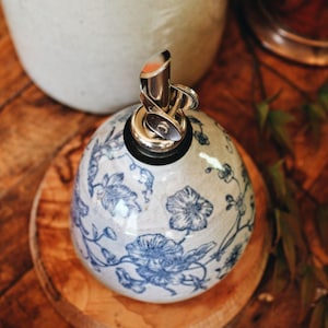 May include: A round ceramic oil dispenser with a glossy off-white finish and a blue floral pattern. The dispenser features a metallic pour spout with a hinged cap and a black rubber seal at the top, providing a functional kitchen accessory.