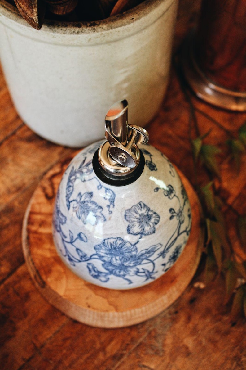 May include: A round ceramic oil dispenser with a glossy off-white finish and a blue floral pattern. The dispenser features a metallic pour spout with a hinged cap and a black rubber seal at the top, providing a functional kitchen accessory.