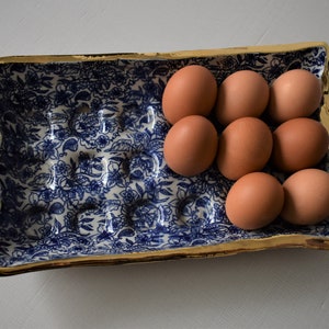 Puede incluir: Un soporte rectangular de cer&aacute;mica para huevos con un estampado floral azul y blanco y un borde dorado. El soporte contiene ocho huevos marrones. El dise&ntilde;o es de estilo cl&aacute;sico y elegante, ideal para la decoraci&oacute;n de la cocina.